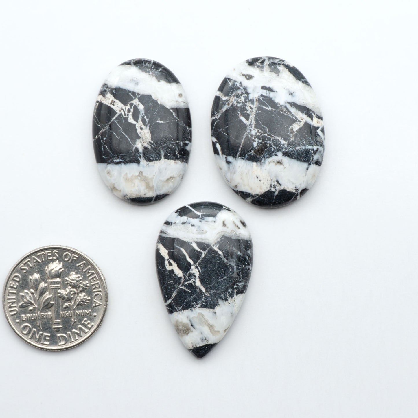 Three black and white marbled stones with a quarter for scale on a white background.Natural White Buffalo Stone Cabochons are semi-precious gemstones cut into shapes ideal for jewelry-making, making them an excellent choice for artisans.