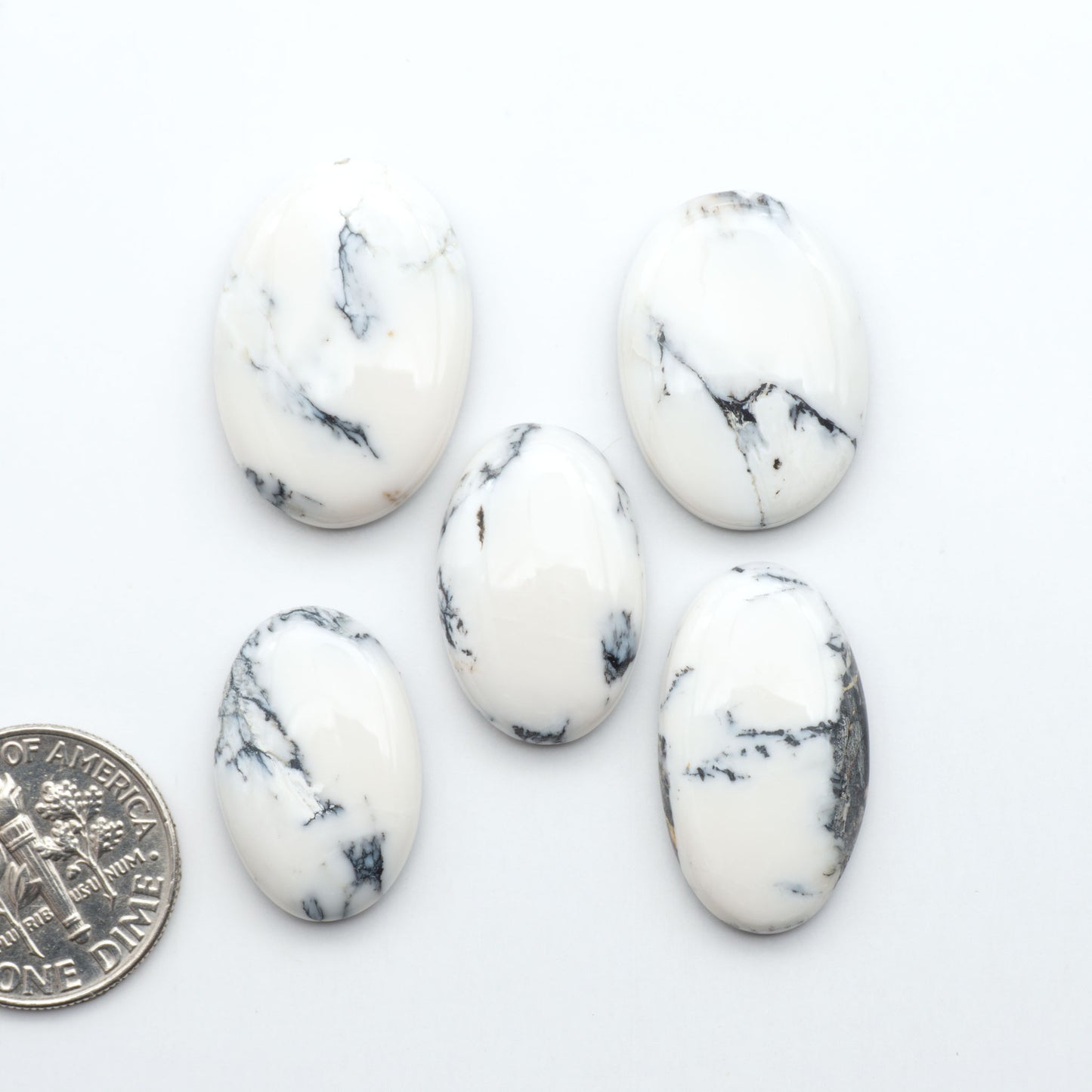 Five white marbled stones with black veins on a white background, one coin for scale.Natural White Buffalo Stone Cabochons are semi-precious gemstones cut into shapes ideal for jewelry-making, making them an excellent choice for artisans.