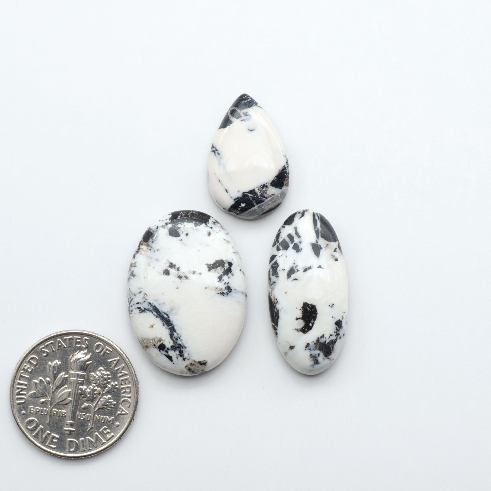 Three oval-shaped white stones with black speckles next to a U.S. dime on a white background.Natural White Buffalo Stone Cabochons are semi-precious gemstones cut into shapes ideal for jewelry-making, making them an excellent choice for artisans.