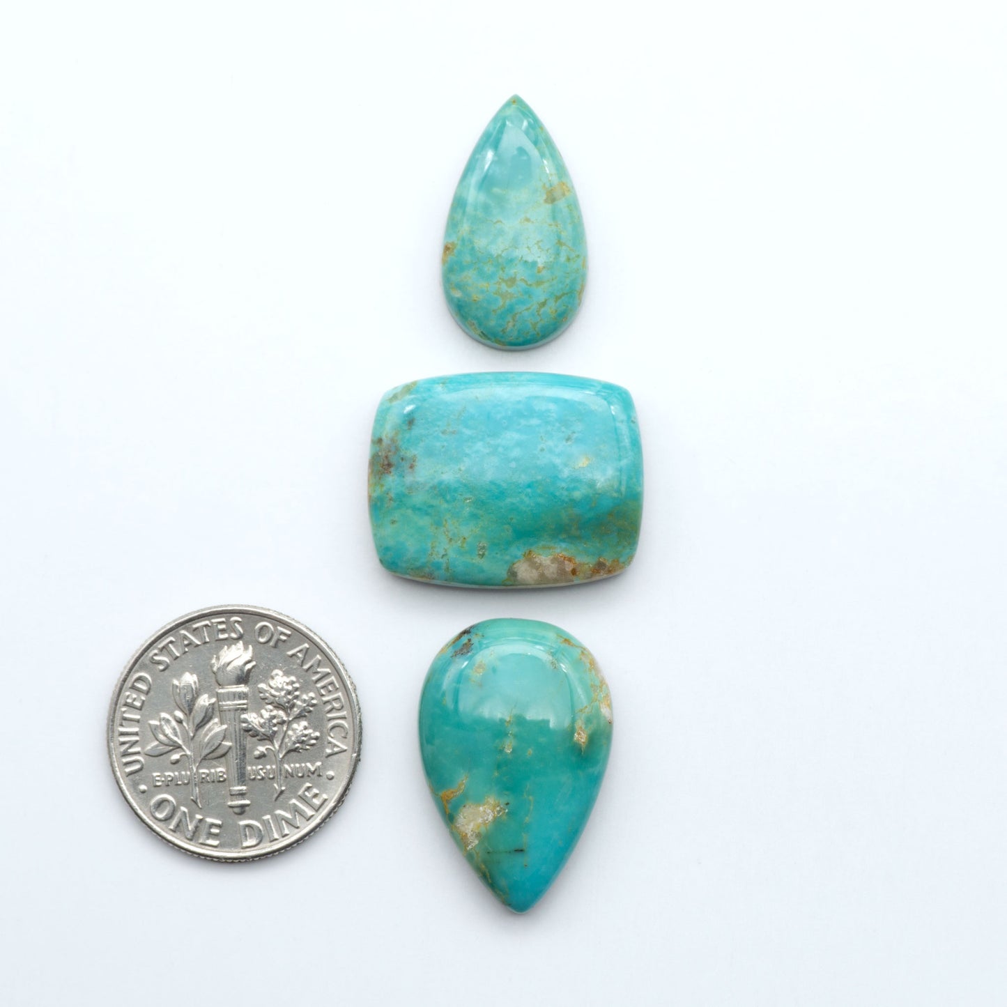 Turquoise stones with a U.S. dime for scale on a white background. Experience the beauty of Turquoise Mountain Cabochons. With their distinctive blue and green color, these gemstones are perfect for any jewelry piece.