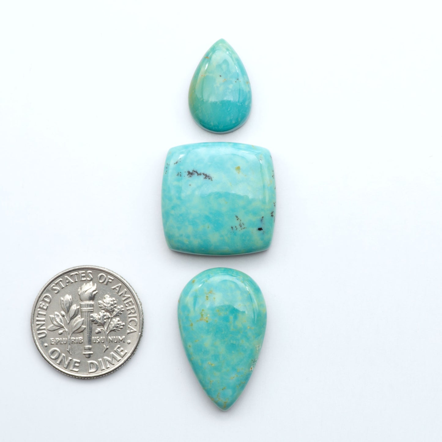 Turquoise stones with a U.S. dime for scale on a white background. Experience the beauty of Turquoise Mountain Cabochons. With their distinctive blue and green color, these gemstones are perfect for any jewelry piece.