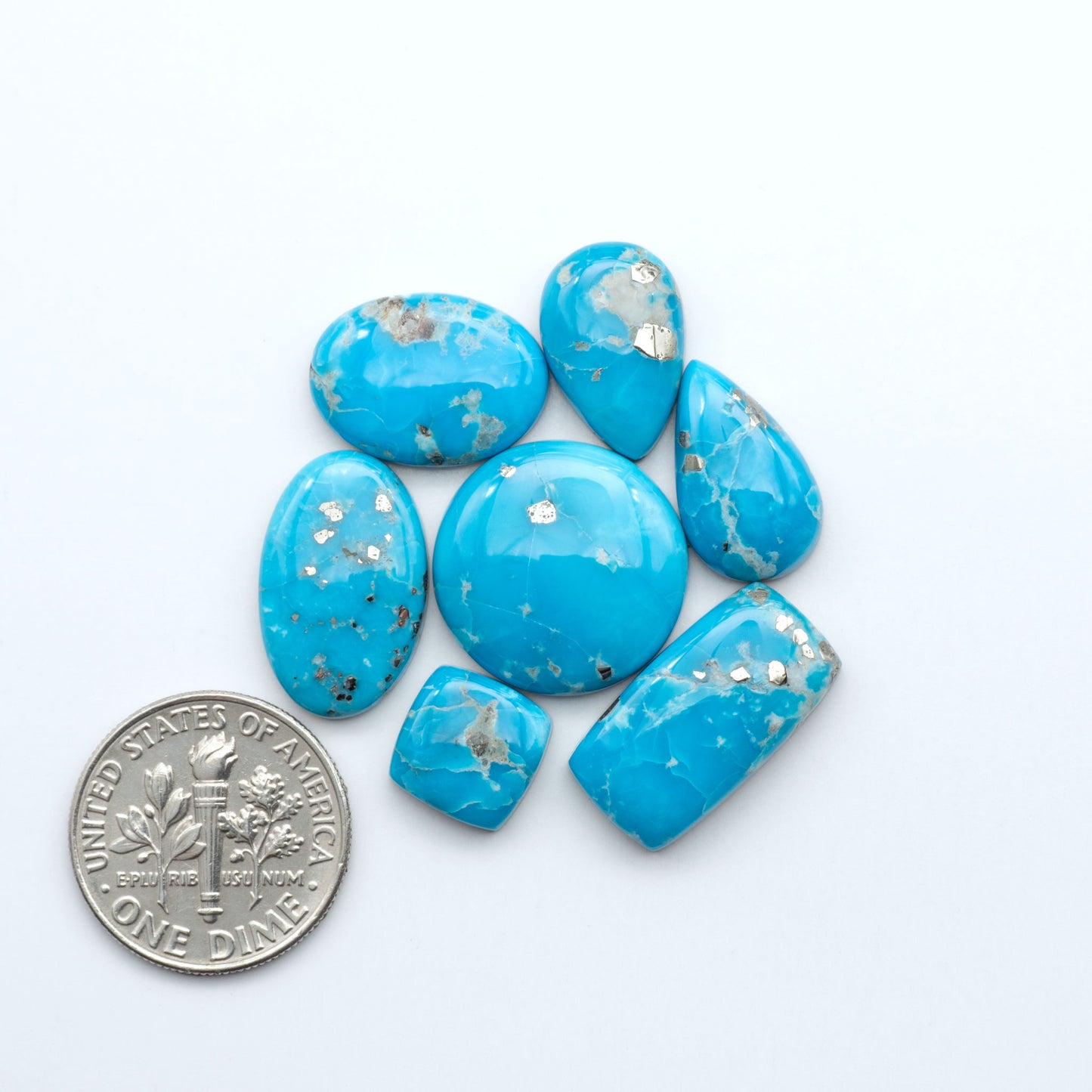 Blue turquoise stones with silver flecks next to a U.S. dime on a white background. Sierra Bella Turquoise cabochons feature rich blue-green color and natural matrix variation. Ideal for silversmiths, jewelers, and custom jewelry designs