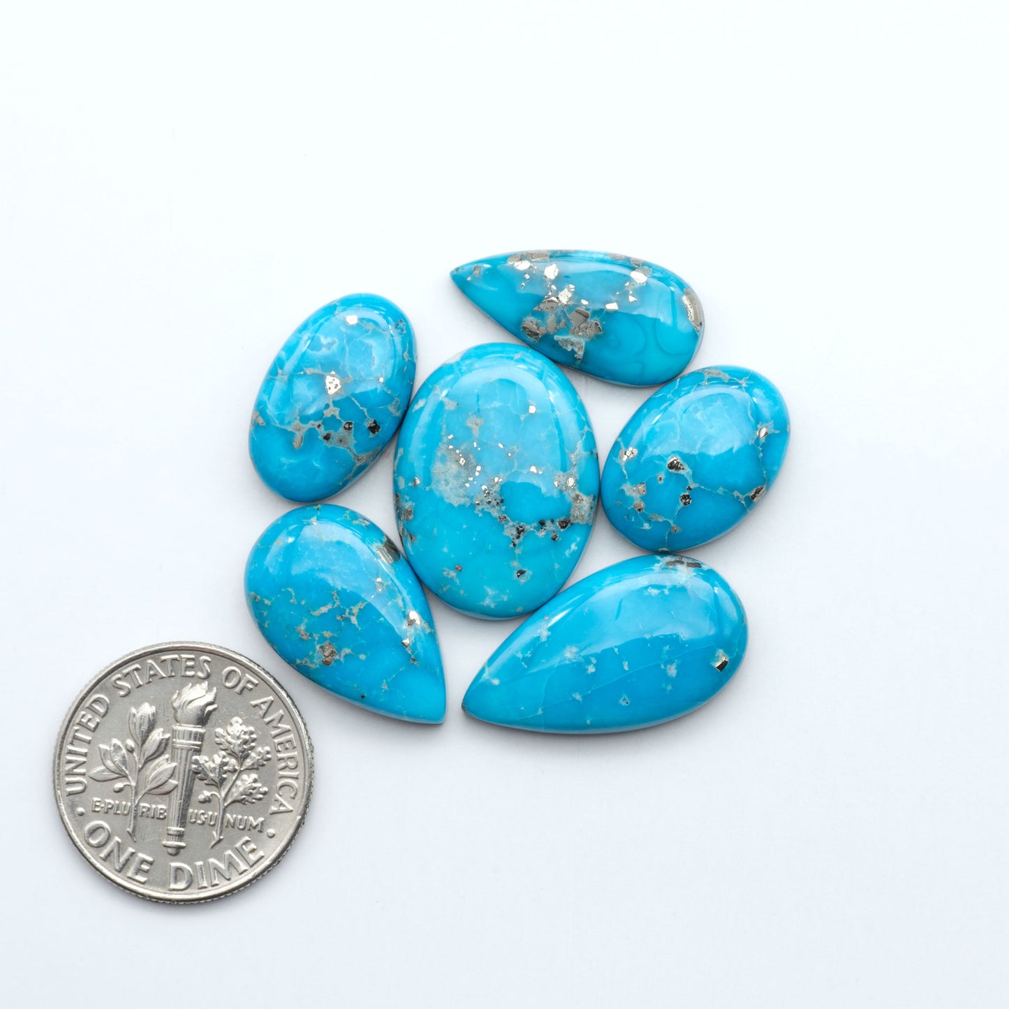 Blue teardrop-shaped stones with gold specks on a white background, next to a U.S. dime for scale. Sierra Bella Turquoise cabochons feature rich blue-green color and natural matrix variation. Ideal for silversmiths, jewelers, and custom jewelry designs