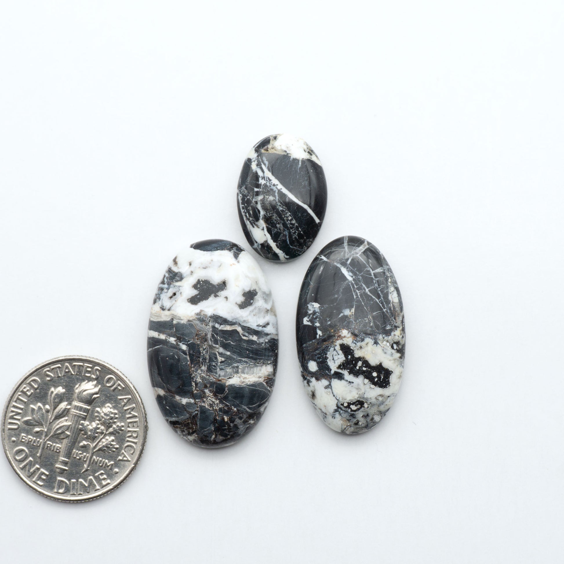 Three oval-shaped black and white marbled stones next to a U.S. dime on a white background.Natural White Buffalo Stone Cabochons are semi-precious gemstones cut into shapes ideal for jewelry-making, making them an excellent choice for artisans.