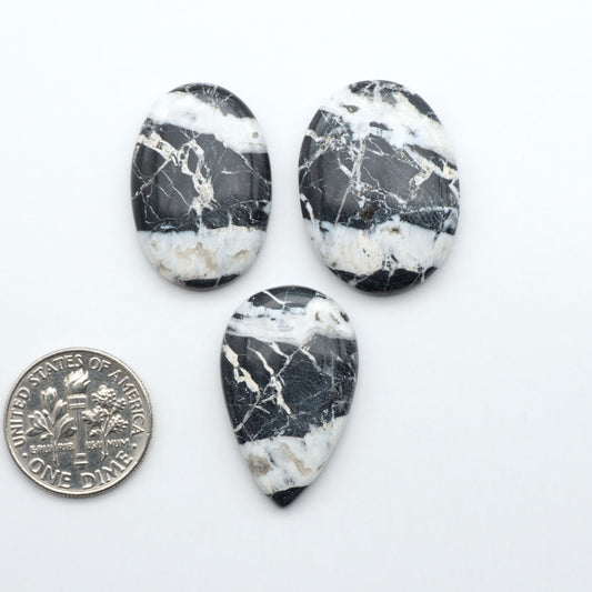 Three black and white marbled stones with a quarter for scale on a white background.Natural White Buffalo Stone Cabochons are semi-precious gemstones cut into shapes ideal for jewelry-making, making them an excellent choice for artisans.