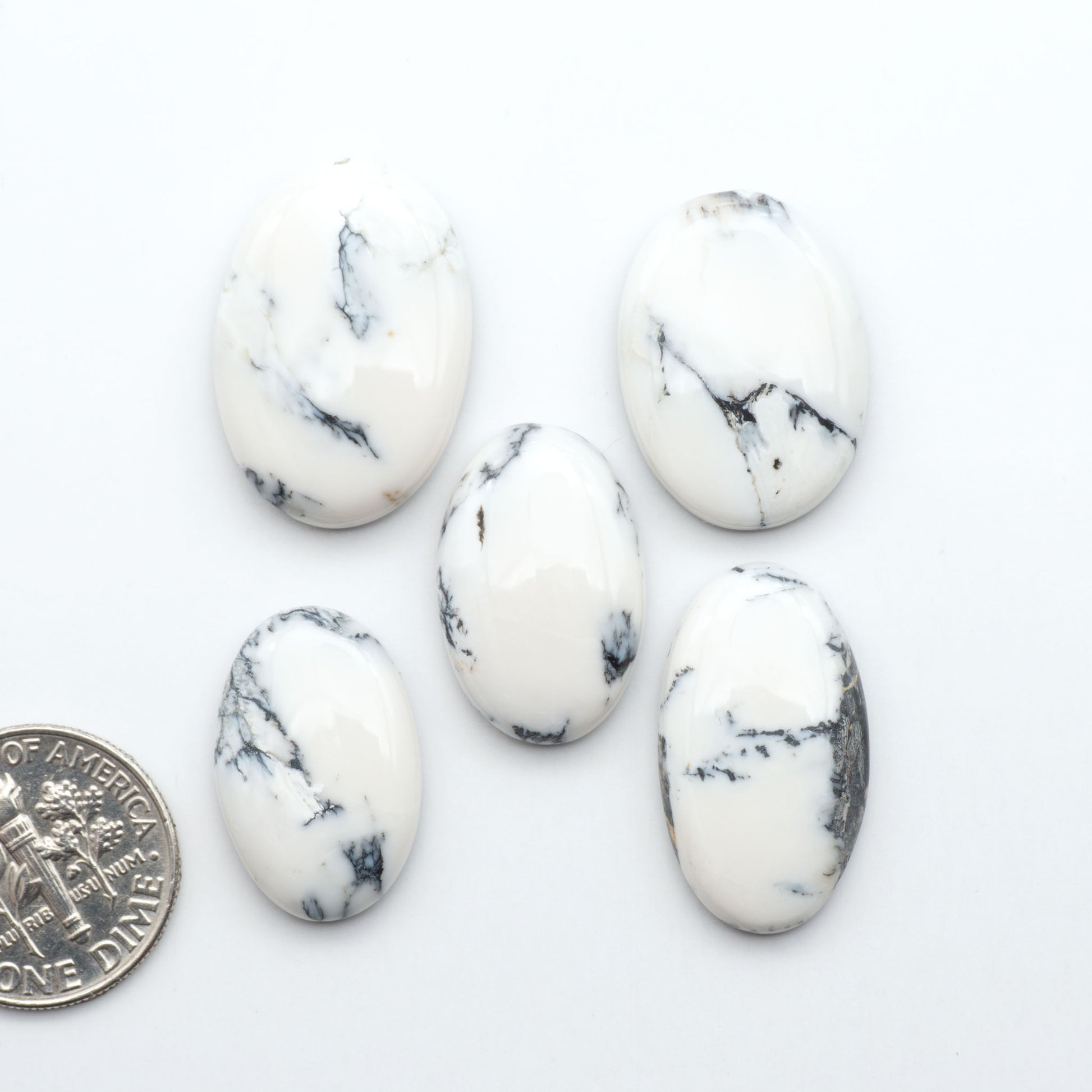 Five white marbled stones with black veins on a white background, one coin for scale.Natural White Buffalo Stone Cabochons are semi-precious gemstones cut into shapes ideal for jewelry-making, making them an excellent choice for artisans.