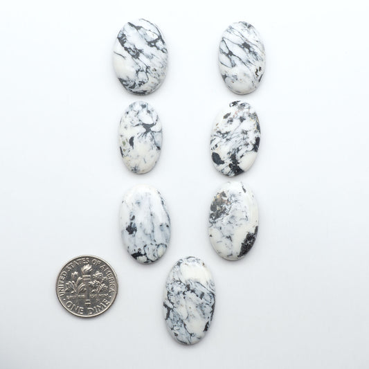 White and black marbled stones with a coin for scale on a white background. Natural White Buffalo Stone Cabochons are semi-precious gemstones cut into shapes ideal for jewelry-making, making them an excellent choice for artisans.