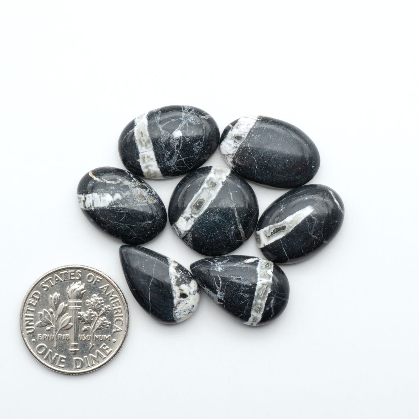 Black and gray marbled stones with silver inclusions on a white background, next to a U.S. dime for scale. Natural White Buffalo Stone Cabochons are semi-precious gemstones cut into shapes ideal for jewelry-making, making them an excellent choice for artisans.
