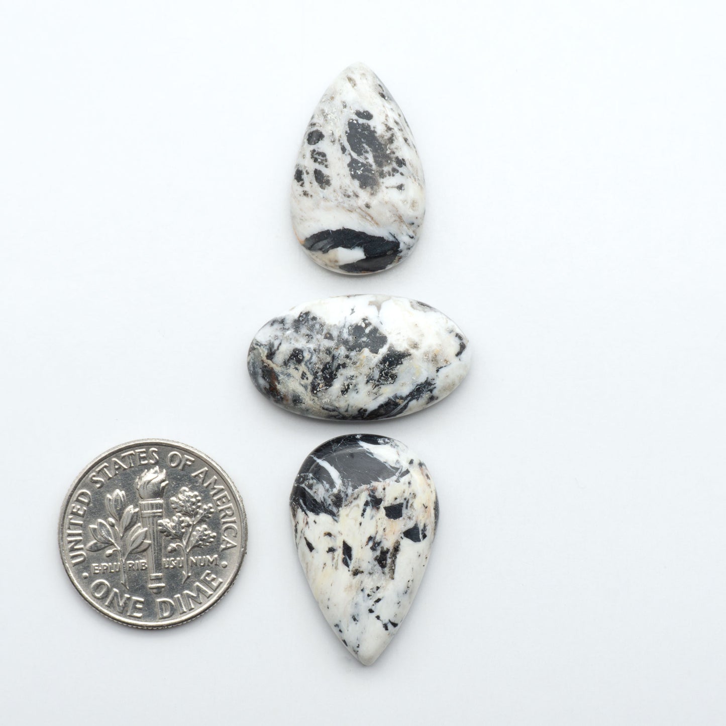 Three teardrop-shaped dalmatian stones on a white background with a dime for scale. Natural White Buffalo Stone Cabochons are semi-precious gemstones cut into shapes ideal for jewelry-making, making them an excellent choice for artisans.
