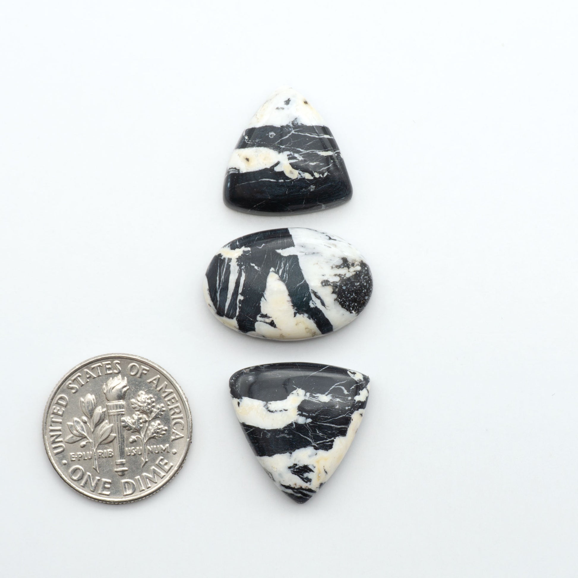 Three black and white marbled stones with a quarter for scale on a white background. Natural White Buffalo Stone Cabochons are semi-precious gemstones cut into shapes ideal for jewelry-making, making them an excellent choice for artisans.