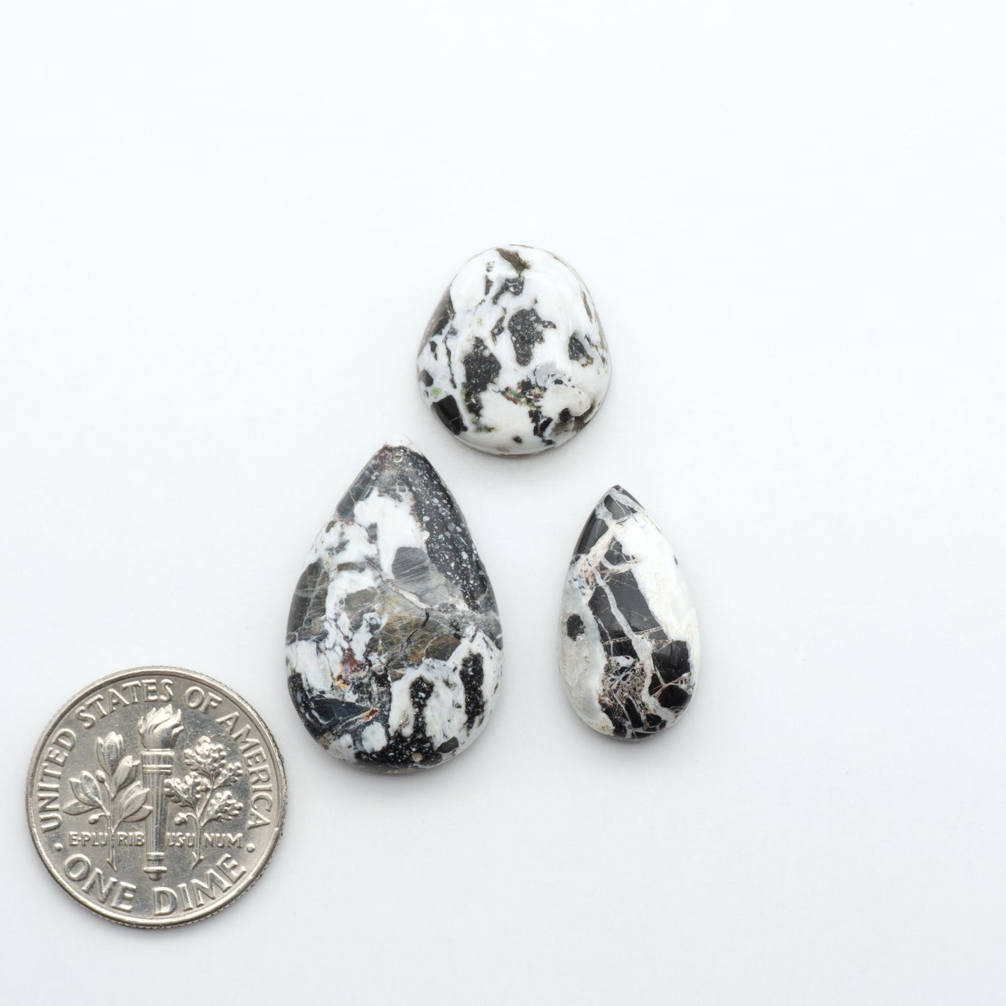 Three teardrop-shaped marbled stones on a white background with a quarter for scale. Natural White Buffalo Stone Cabochons are semi-precious gemstones cut into shapes ideal for jewelry-making, making them an excellent choice for artisans.