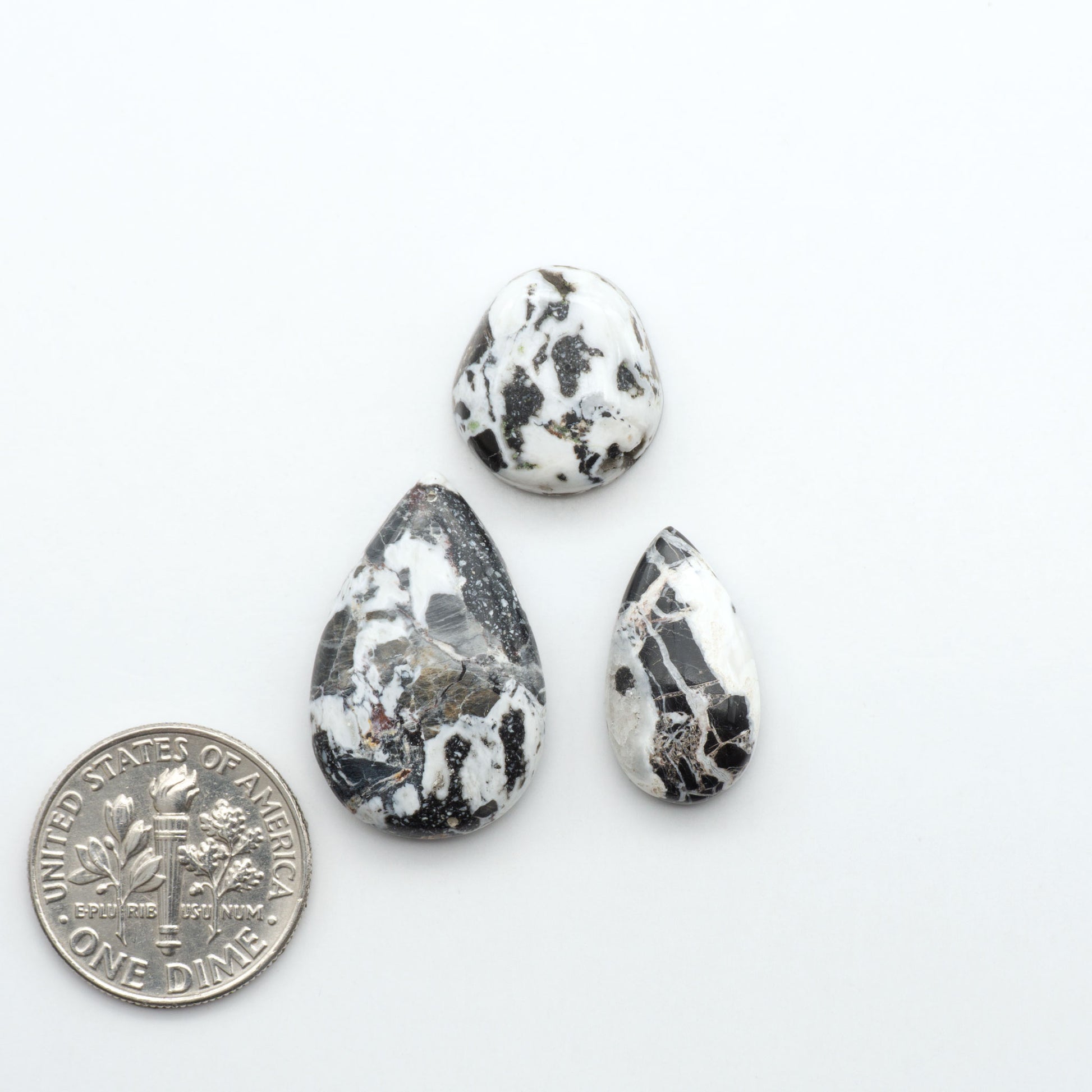 Three teardrop-shaped marbled stones on a white background with a quarter for scale. Natural White Buffalo Stone Cabochons are semi-precious gemstones cut into shapes ideal for jewelry-making, making them an excellent choice for artisans.