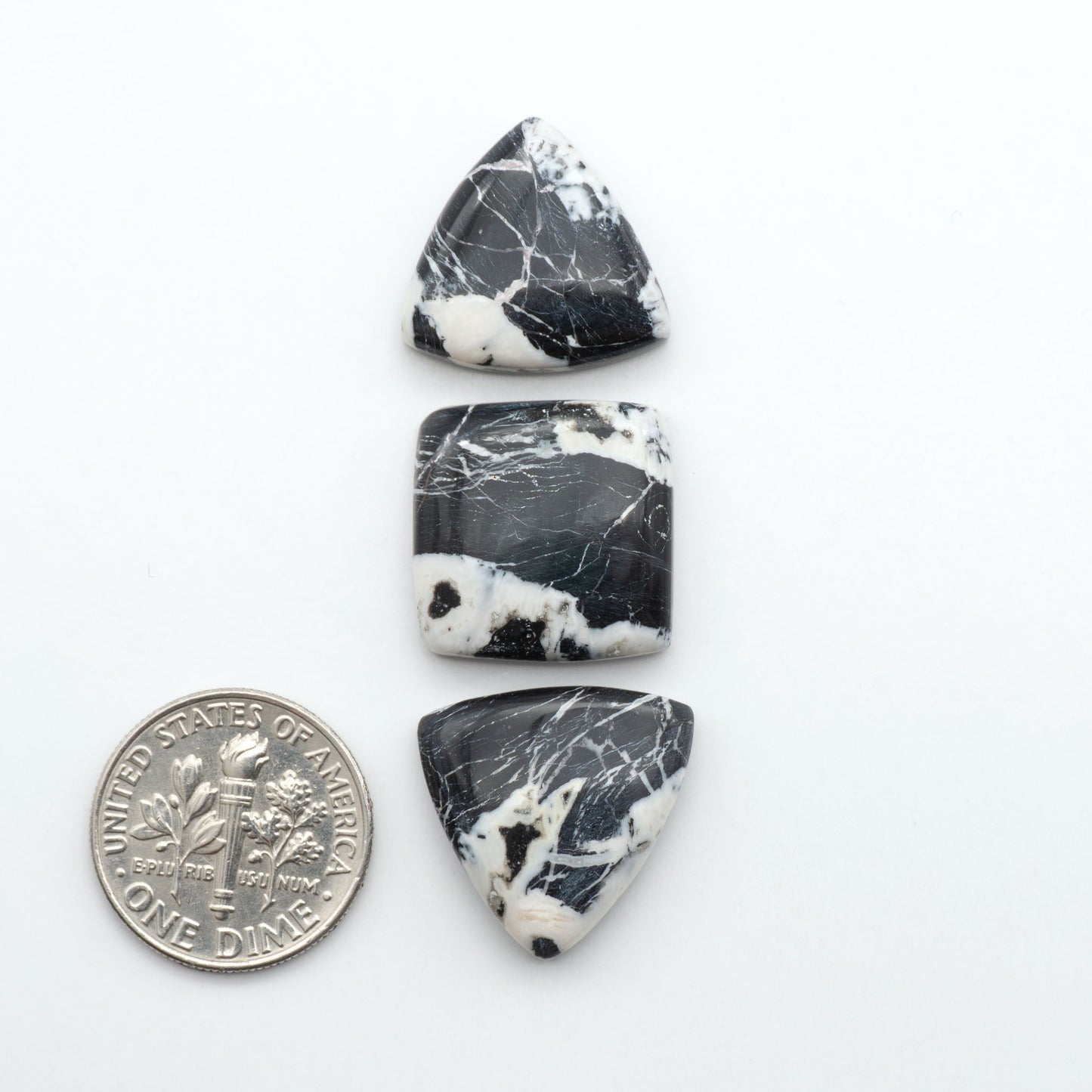Three black and white marbled stones with a quarter for scale on a white background. Natural White Buffalo Stone Cabochons are semi-precious gemstones cut into shapes ideal for jewelry-making, making them an excellent choice for artisans.