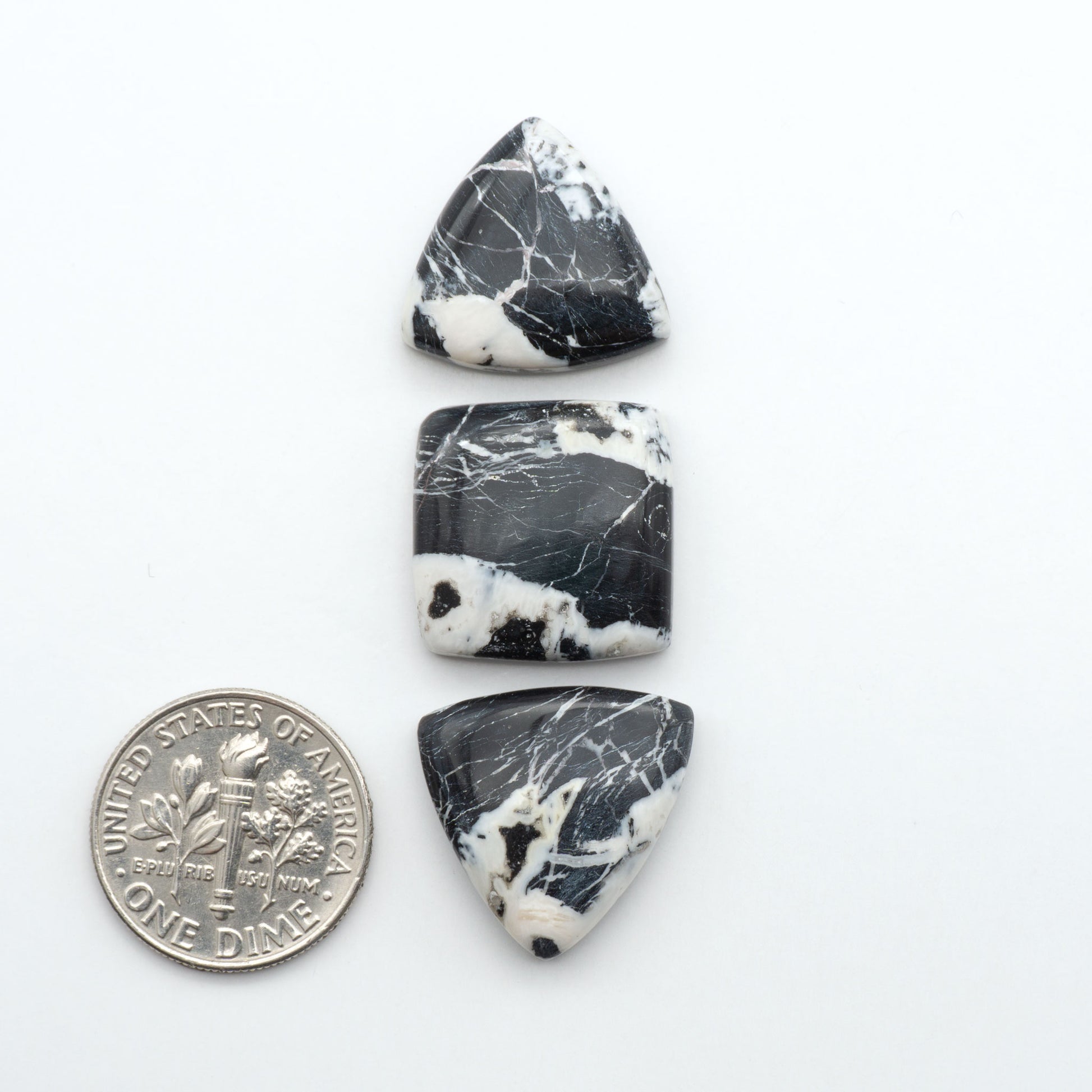Three black and white marbled stones with a quarter for scale on a white background. Natural White Buffalo Stone Cabochons are semi-precious gemstones cut into shapes ideal for jewelry-making, making them an excellent choice for artisans.
