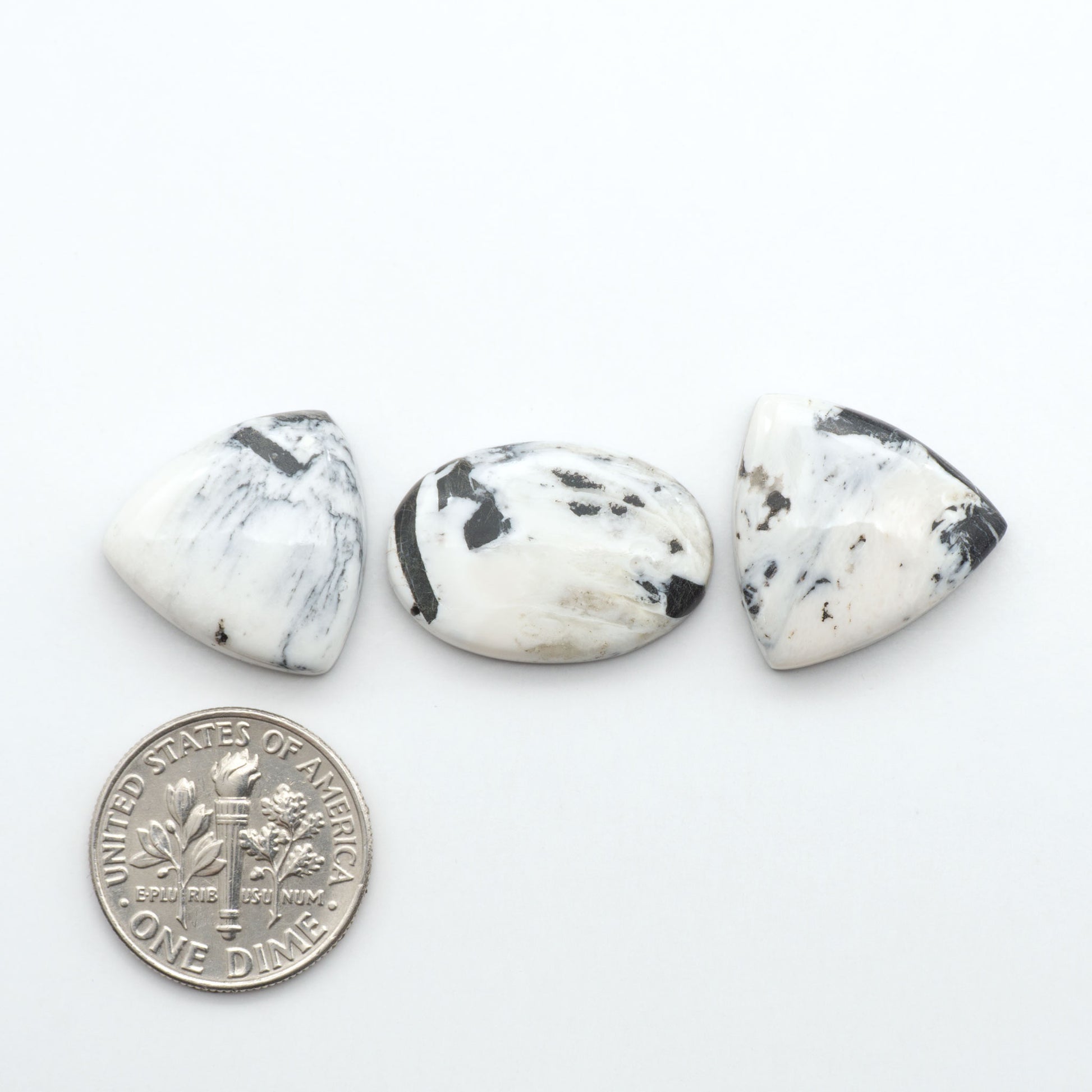 Three white marbled stones with black spots on a white background, next to a U.S. dime for scale. Natural White Buffalo Stone Cabochons are semi-precious gemstones cut into shapes ideal for jewelry-making, making them an excellent choice for artisans.