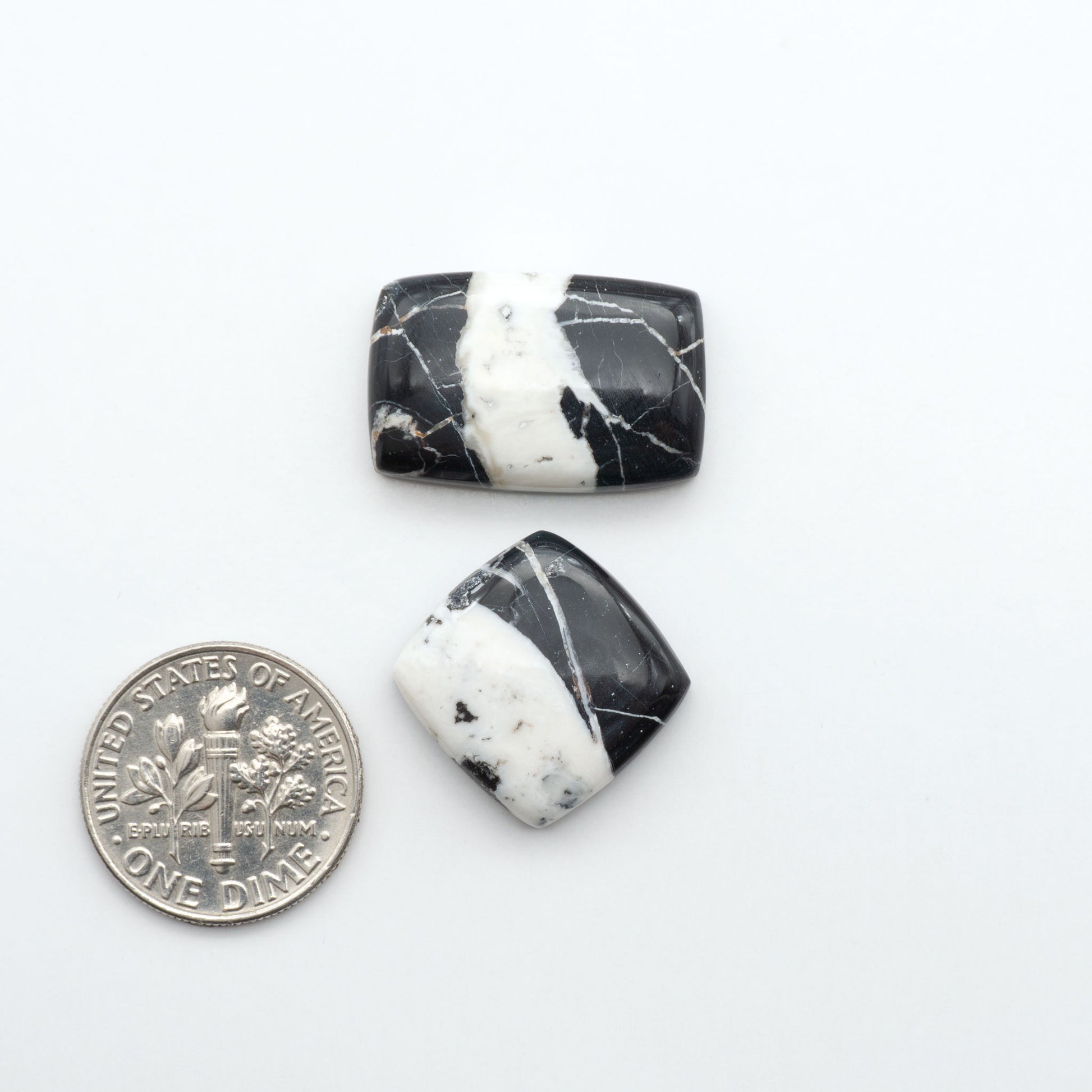 Two black and white marbled stones next to a U.S. dime on a white background. Natural White Buffalo Stone Cabochons are semi-precious gemstones cut into shapes ideal for jewelry-making, making them an excellent choice for artisans.