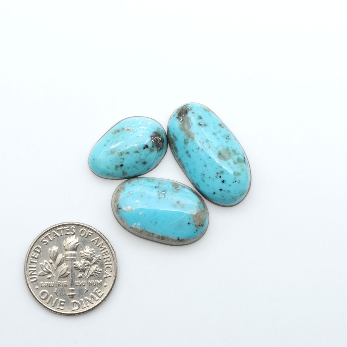 Nacozari Turquoise cabochons, blue to greenish-blue gemstones with black or brown matrix and pyrite on a white background, with a US dime for scale